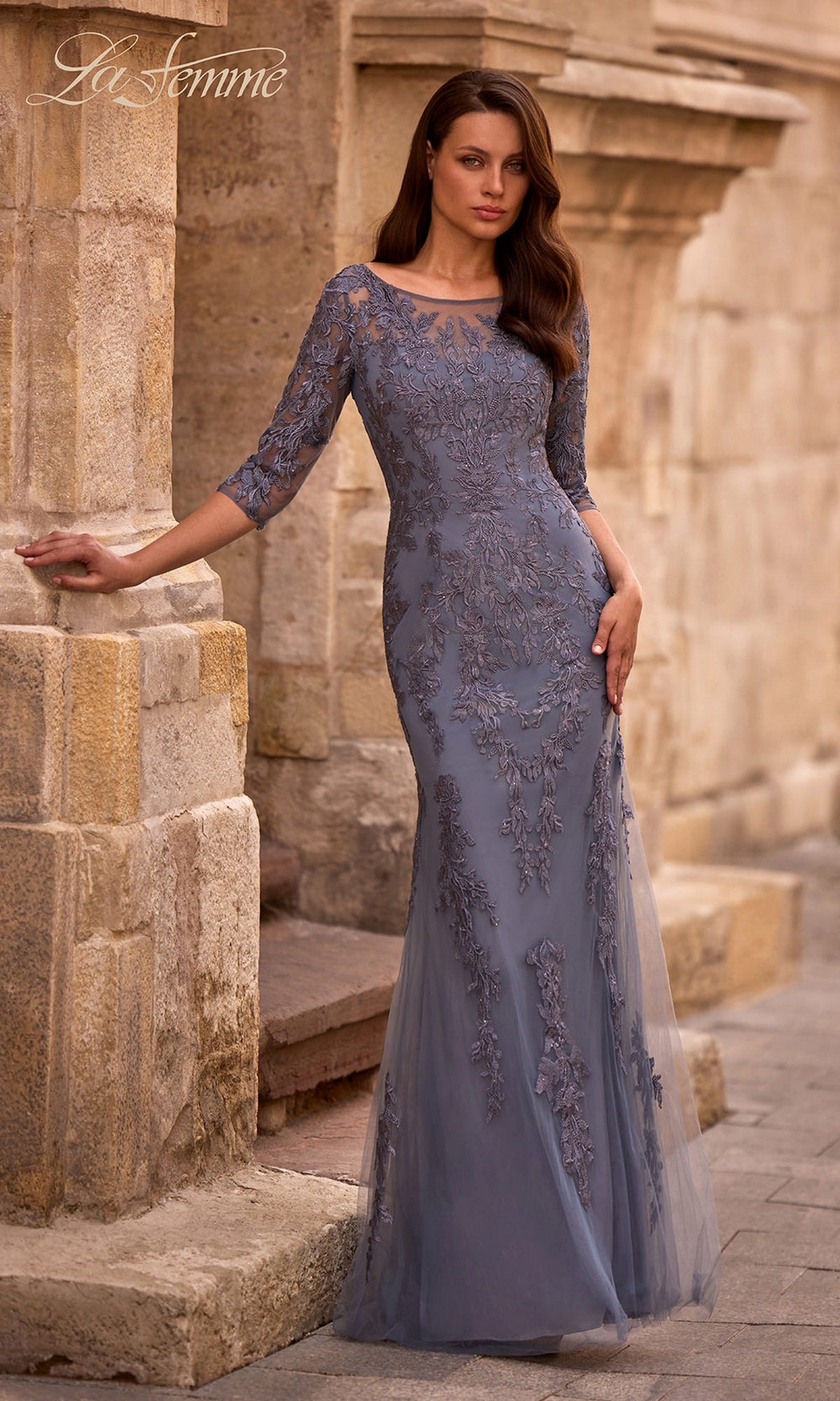 A woman in the slate Long Formal Dress: La Femme 29226 with lace embroidery and three-quarter sleeves stands by a stone wall outdoors.